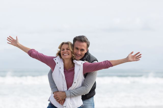 Mature couple enjoying a joyful moment on the beach, with the man embracing the woman from behind as she stretches her arms out. Ideal for use in advertisements, travel brochures, romantic getaway promotions, and lifestyle blogs focusing on relationships, happiness, and outdoor activities.