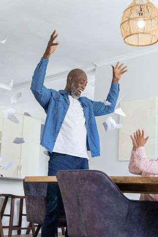 Joyful senior couple celebrating at home, man raising arms amid flying papers