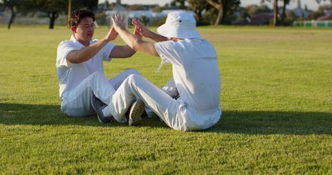 Two teammates sitting on grass clapping in white cricket kit celebrating partnership