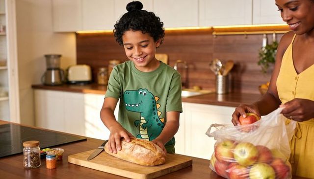 Smiling child slicing crusty bread at kitchen island while adult holding bag of apples