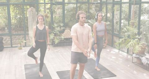 Group practicing yoga inside glass greenhouse wearing headphones balancing on mats