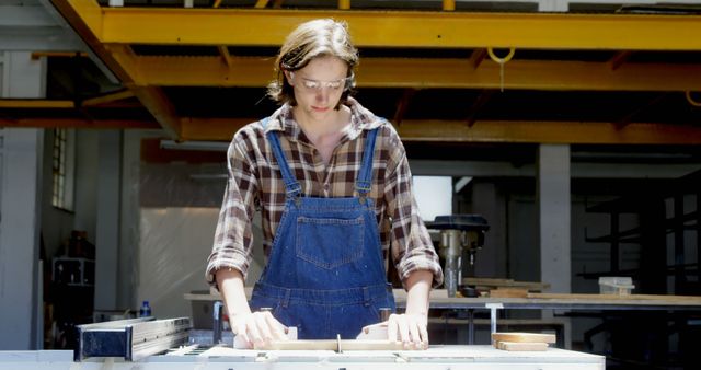 Young adult carpenter wearing safety glasses, working with wood in a workshop on sunny day. Suggested use: articles on woodworking, safety in carpentry, craftsmanship, or vocational training.