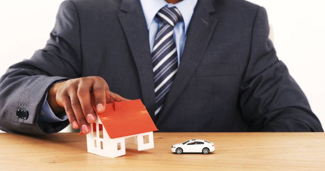 An African American businessman is presenting a small model house and a toy car on a desk, with copy space. It symbolizes concepts related to real estate, property ownership, or financial planning.