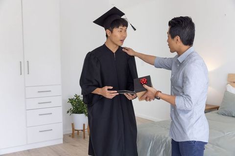 Asian graduate celebrating wearing cap gown holding diploma as friend presenting gift