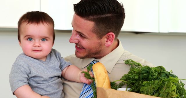 Father holding his baby son and paper bag of groceries at home in the kitchen