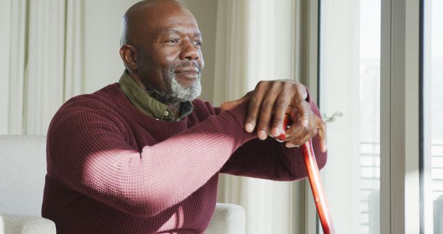 An elderly man is sitting near a window with his hands resting on his walking cane, looking thoughtful and relaxed. Perfect for articles on aging, senior lifestyle, or healthcare for older adults. Ideal for magazines, blogs, and advertisements relating to retirement, mental health, or understanding the inner thoughts of senior citizens.