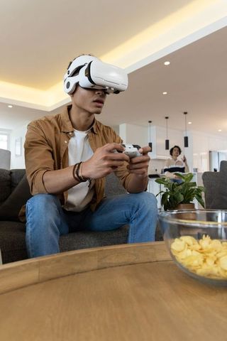 Asian man wearing vr headset playing with controller on sofa in modern lounge while friend watching
