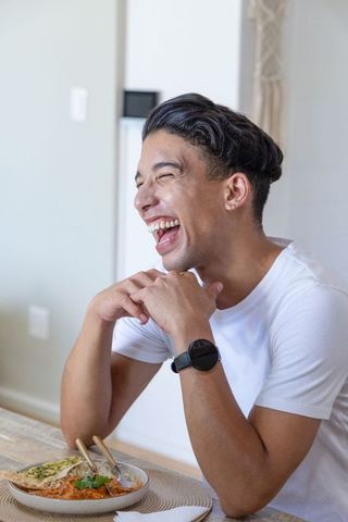 Laughing african american man enjoying meal at dining table, white tee, smartwatch, candid