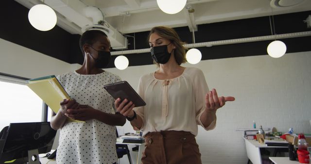 Two professional women discuss work in a modern office while wearing masks. One holds a tablet, the other holds folders, emphasizing teamwork and collaboration. This image is useful for articles on professional working environments, safety measures during a pandemic, or corporate communication. Ideal for business, health, and workplace safety themes.