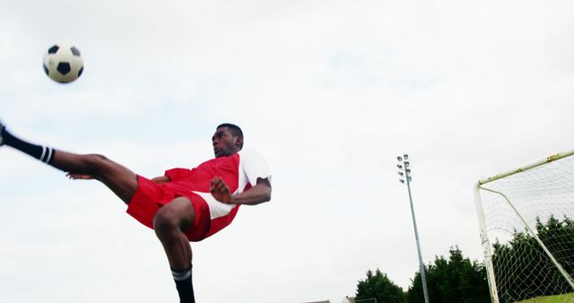 A young African American male athlete in a red soccer uniform performs a high kick to control the ball during a game, with copy space. His dynamic action captures the intensity and skill involved in the sport of soccer.