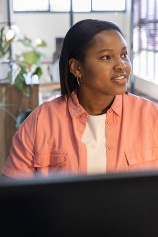 African american woman working at modern desk looking thoughtful in sunlit office near blinds