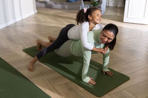 Diverse mother practicing plank while joyful daughter rides on back on green yoga mat