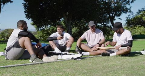 Diverse friends relaxing after cricket practice, sitting cross-legged on sunny park grass