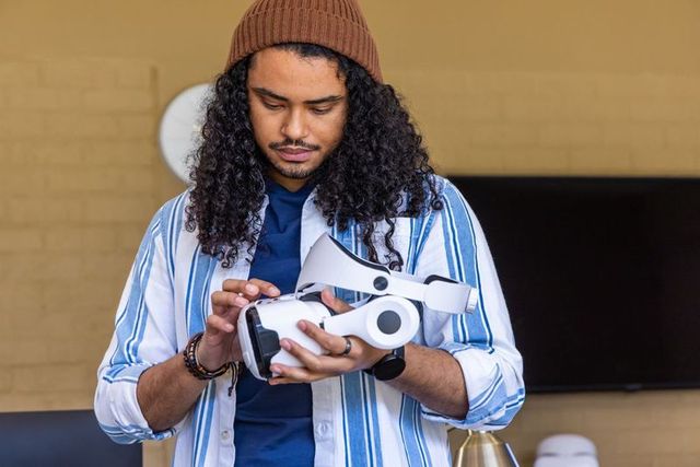 Young man inspecting white vr headset in modern lounge wearing beanie and striped shirt