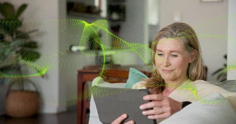 Mature woman holding tablet and relaxing on sofa with neon green data waveform overlay