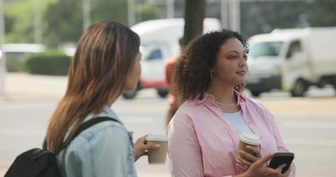 Two women chatting on city curb holding takeaway coffee cups and checking smartphone