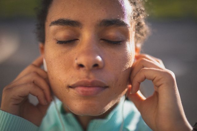 This image depicts a biracial woman with her eyes closed, putting earphones in her ears before starting her outdoor exercise routine. She appears calm and focused, embodying a healthy lifestyle and self-care. This image is perfect for use in fitness blogs, health and wellness websites, or advertisements promoting outdoor activities and exercise gear.