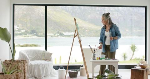 Mature woman painting at easel by lakeside window overlooking mountains holding brushes