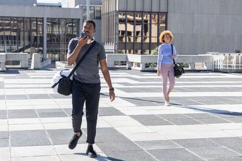 African american couple walking urban rooftop plaza carrying gym bags and using smartphone