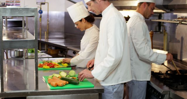 Professional chefs are wearing white chef uniforms while preparing meals in a restaurant kitchen. Various fresh vegetables like carrots, zucchinis, and peppers are placed on green cutting boards. This photo can be used to represent teamwork, culinary expertise, and the hard work that goes into restaurant meal preparation. Ideal for articles, advertisements, and materials related to gastronomy, restaurant management, or culinary training.