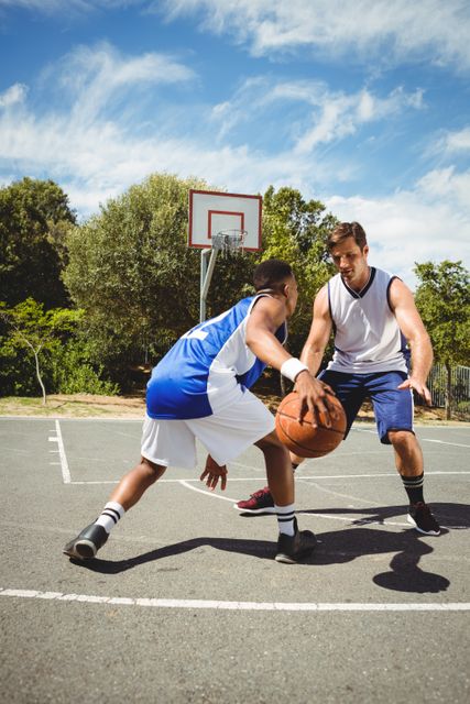 Two basketball players are practicing on an outdoor court on a sunny day. One player is dribbling the ball while the other is defending. This image can be used for sports training materials, fitness and exercise promotions, teamwork and competition concepts, and recreational activity advertisements.