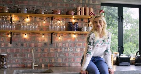 Young woman sitting on kitchen counter wearing cozy sweater with string lights, rustic brick