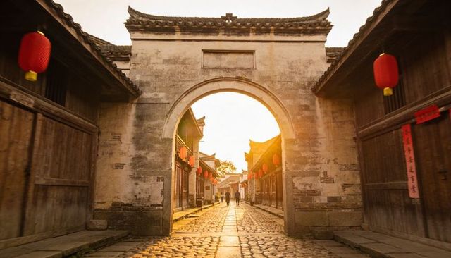 Sunlit stone archway leading cobblestone lane with red lanterns in historic chinese village