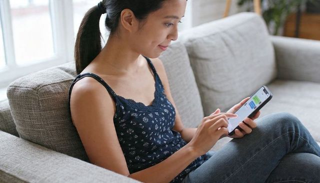 Asian woman using smartphone on sofa at home, texting and scrolling in cozy sunlight