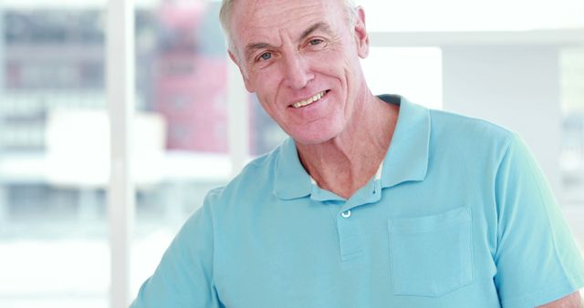 Senior man in blue polo shirt is smiling indoors with natural light coming through windows in background. Set in a bright, modern environment, his relaxed and confident demeanor emphasizes positivity and well-being. This image can be used in advertisements, healthcare promotions, elder care services, or lifestyle articles to convey a friendly and approachable atmosphere.