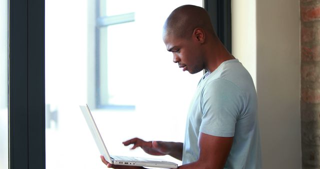 Young Man Working on Laptop by Window in Casual Clothing - Download Free Stock Images Pikwizard.com