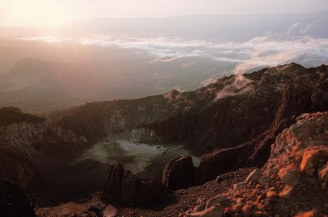 This image captures a dramatic view of a volcanic crater bathed in the warm light of sunset. The rugged landscape contrasts with the soft clouds hovering above, creating a breathtaking scene that exudes a sense of adventure and exploration. Ideal for travel agencies, adventure blogs, or nature magazines, this image could be used to inspire travel, promote eco-tourism, or highlight geological features.