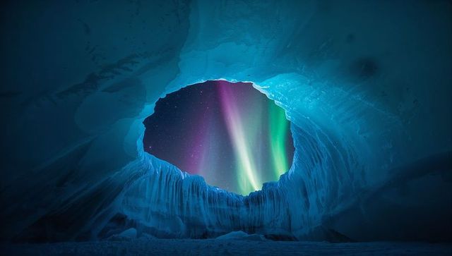 Framing oval ice cave portal revealing vibrant aurora borealis over frozen glacier floor