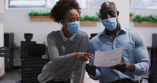 Two colleagues wearing protective face masks working together in a modern office setting. They are discussing a document, demonstrating teamwork and cooperation. This visual is poised to be informative in business and professional presentations, articles about workplace safety during the COVID-19 pandemic, teamwork, and collaboration.