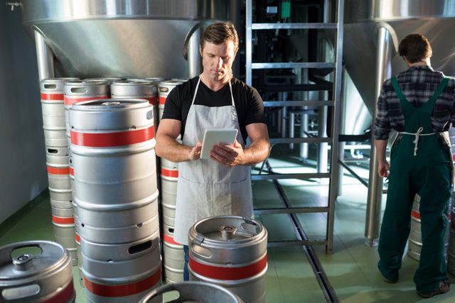 Brewery workers managing beer kegs in an industrial setting. One worker is using a tablet to check inventory or production data, while another is working in the background. This image can be used for articles or advertisements related to brewing, beer production, industrial work, or technology in manufacturing.