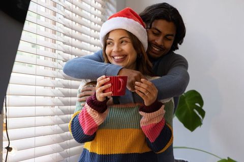 Diverse couple hugging at home wearing santa hat and colorful sweaters holding red mug
