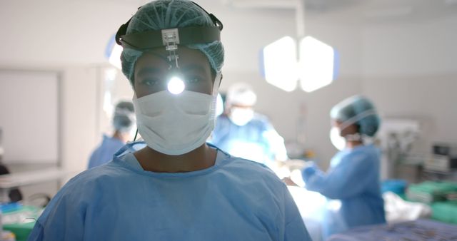 Surgeon wearing scrubs, mask, and headlamp, standing in an operating room with focused medical team working in the background. Can be used for healthcare promotions, medical articles, hospital brochures, surgery-related content, or illustrating surgical procedures in medical publications.