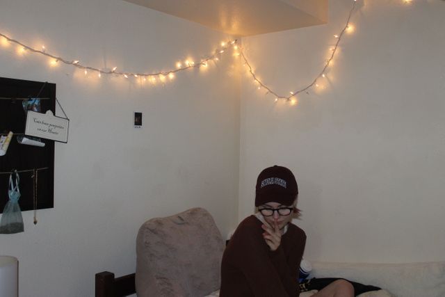 Teen sitting in a bedroom adorned with fairy lights, creating a relaxing atmosphere. Wearing a burgundy hat and glasses, providing a cozy vibes. Ideal for content related to home decor, teen lifestyle, and relaxation tips.