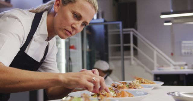 This stock photo shows a professional chef meticulously plating gourmet dishes in a modern restaurant kitchen. Perfect for use in articles or marketing materials related to fine dining, culinary arts, professional cooking, or restaurant quality standards. Ideal for illustrating tutorials on food presentation or showcasing the dedication and precision required in professional cooking.