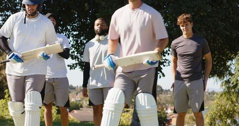 Multiracial cricket team walking together on sunny field carrying bats and pads