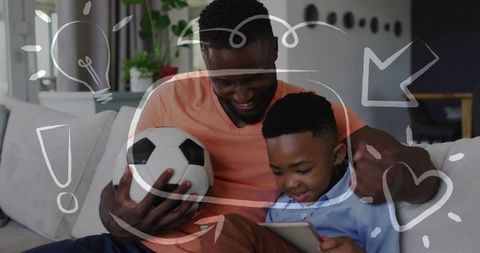 Father and son bonding on couch using tablet while holding soccer ball and smiling
