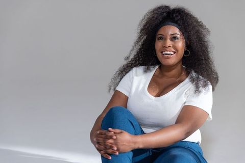 Smiling african american woman sitting with knee up in studio wearing white top and jeans curly hair