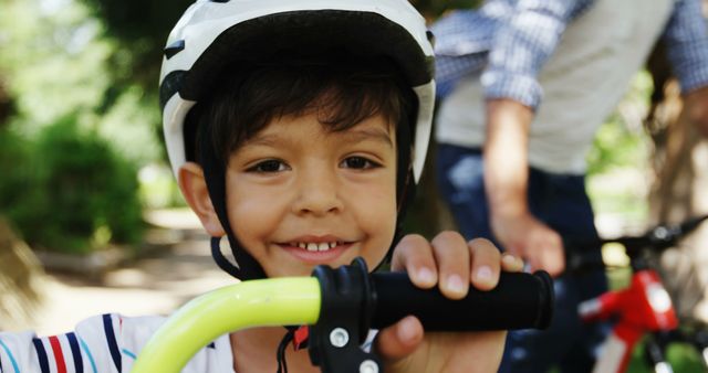 Young boy smiling while holding bicycle handlebars in outdoor park. Perfect for activities involving children's safety gear, promoting outdoor play, and cycling safety tips.