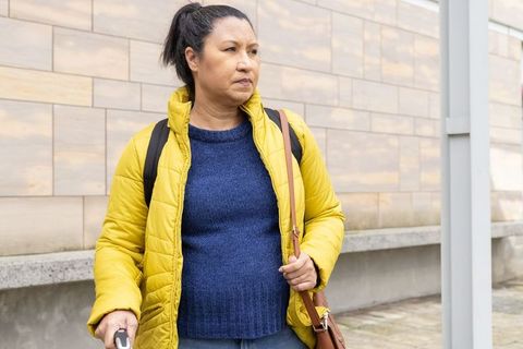 Mature woman standing holding white cane and shoulder bag wearing yellow puffer jacket