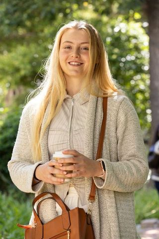 Smiling woman holding takeaway coffee on sunlit park path wearing knit cardigan, tan bag