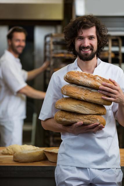 Male baker smiling and carrying a stack of freshly baked breads in a professional kitchen. Another baker working in the background. Can be used for themes related to baking, culinary arts, professional kitchens, or artisan bread making.