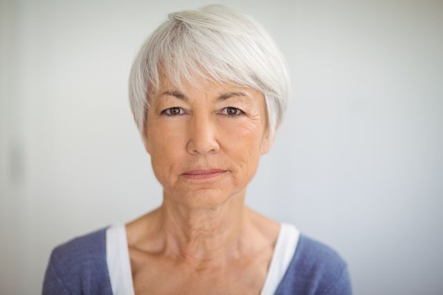 This image depicts a confident senior woman with gray hair, looking directly at the camera with a serious expression. The natural light and home setting add a sense of comfort and authenticity. This image can be used in articles or advertisements related to aging, senior lifestyle, healthcare, retirement planning, and personal stories of elderly individuals.