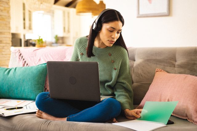 Biracial woman working on laptop in cozy living room, wearing headphones and casual attire. Ideal for illustrating remote work, home office setups, modern lifestyle, and productivity. Perfect for blogs, articles, and advertisements related to working from home, technology, and comfortable living.