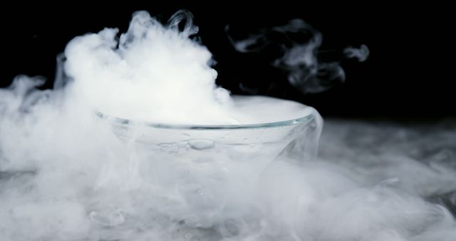 A glass bowl is enveloped in a cloud of white smoke or vapor, from dry ice, creating a mysterious and dramatic effect. The smoke billows around the transparent container, emphasizing the contrast between the clear glass and the dense fog.
