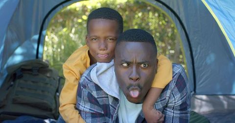 African american father and son making funny faces in tent while camping together