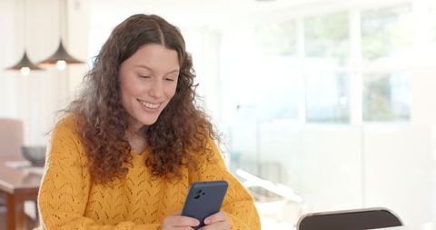 Smiling woman texting on smartphone in sunlit modern home wearing mustard-yellow sweater